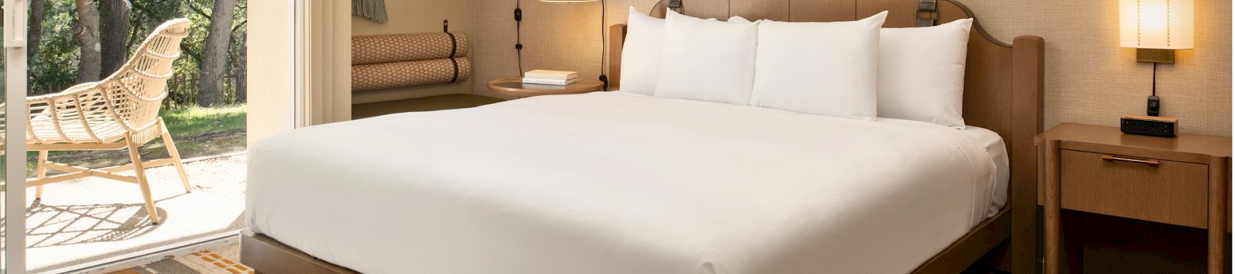 A bright hotel bedroom with a large bed, bedside lamps, patterned rug, and an open window showing trees outside.