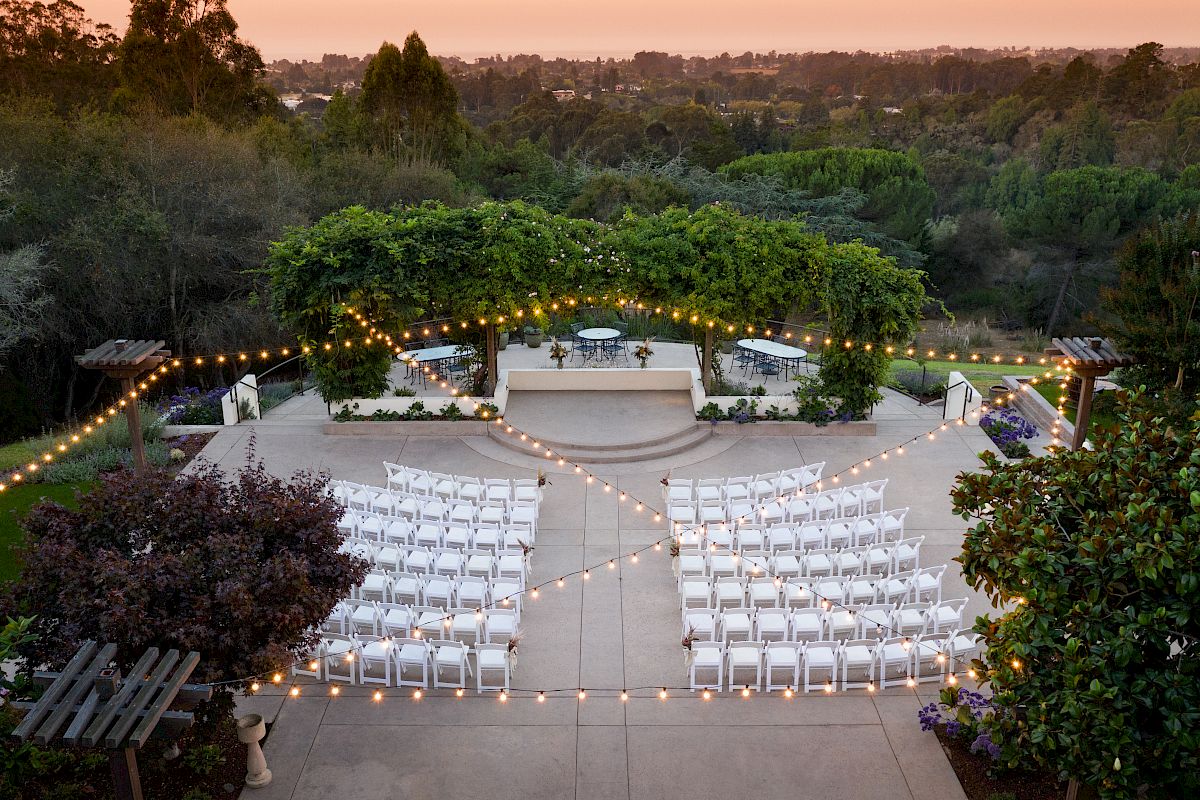 An outdoor venue with white chairs set up for an event, surrounded by string lights and greenery, under a cloudy sunrise or sunset sky.